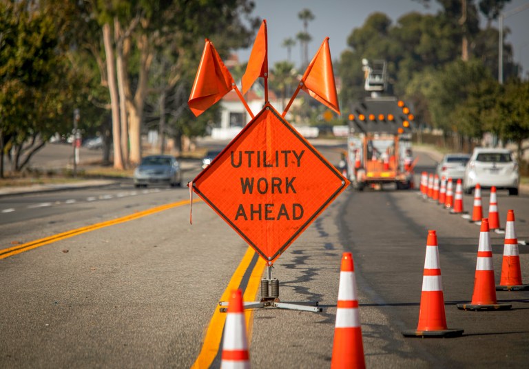 Traffic sign with flags reading "Utility Work Ahead" with traffic cones on road with electronic arrow pointing to the right to divert traffic